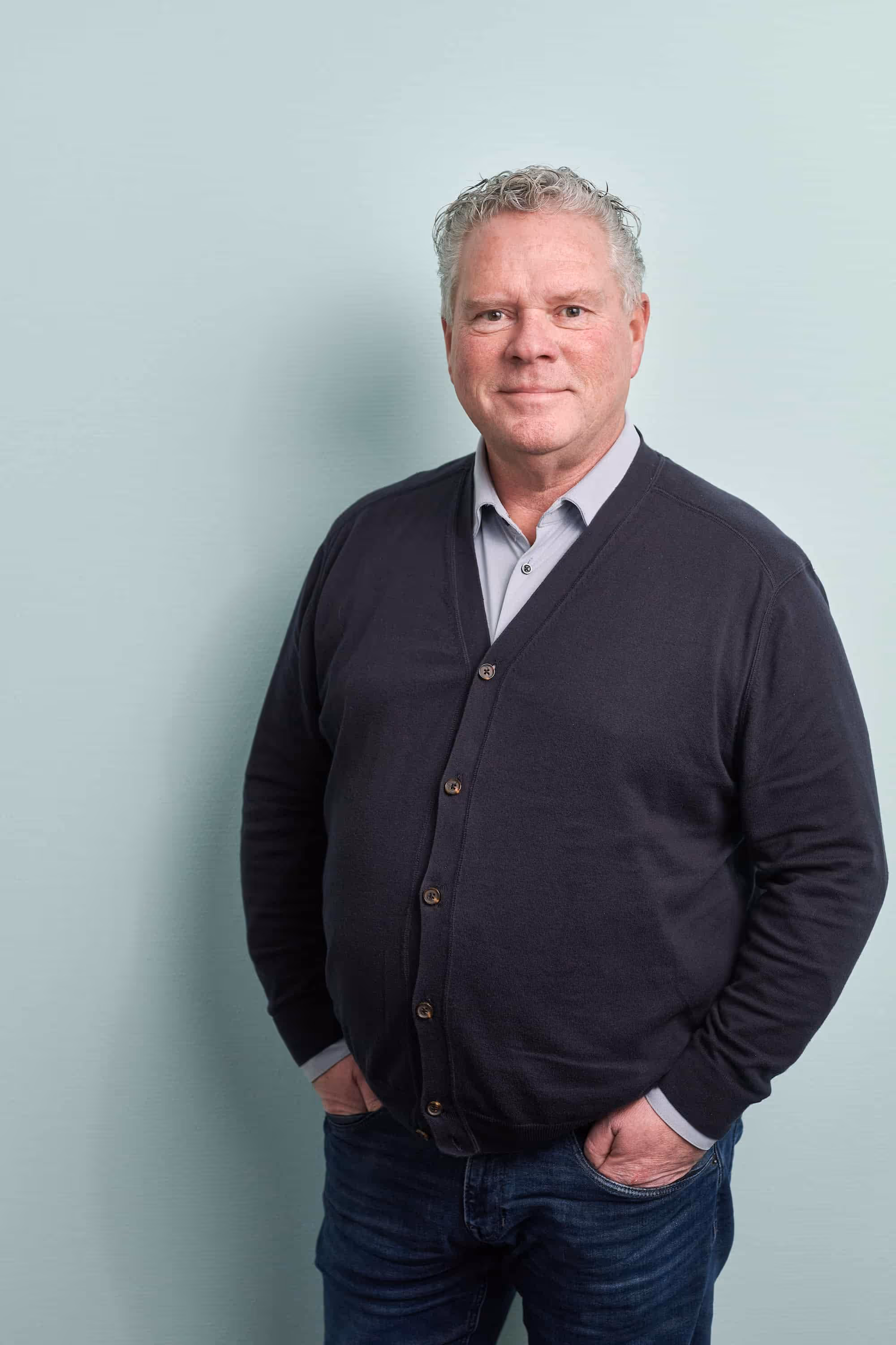 Middelbare man met grijs golvend haar, grijze overhemd en een donkergrijze vest, staand met handen in zijn broekzakken tegen een lichtblauwe achtergrond.