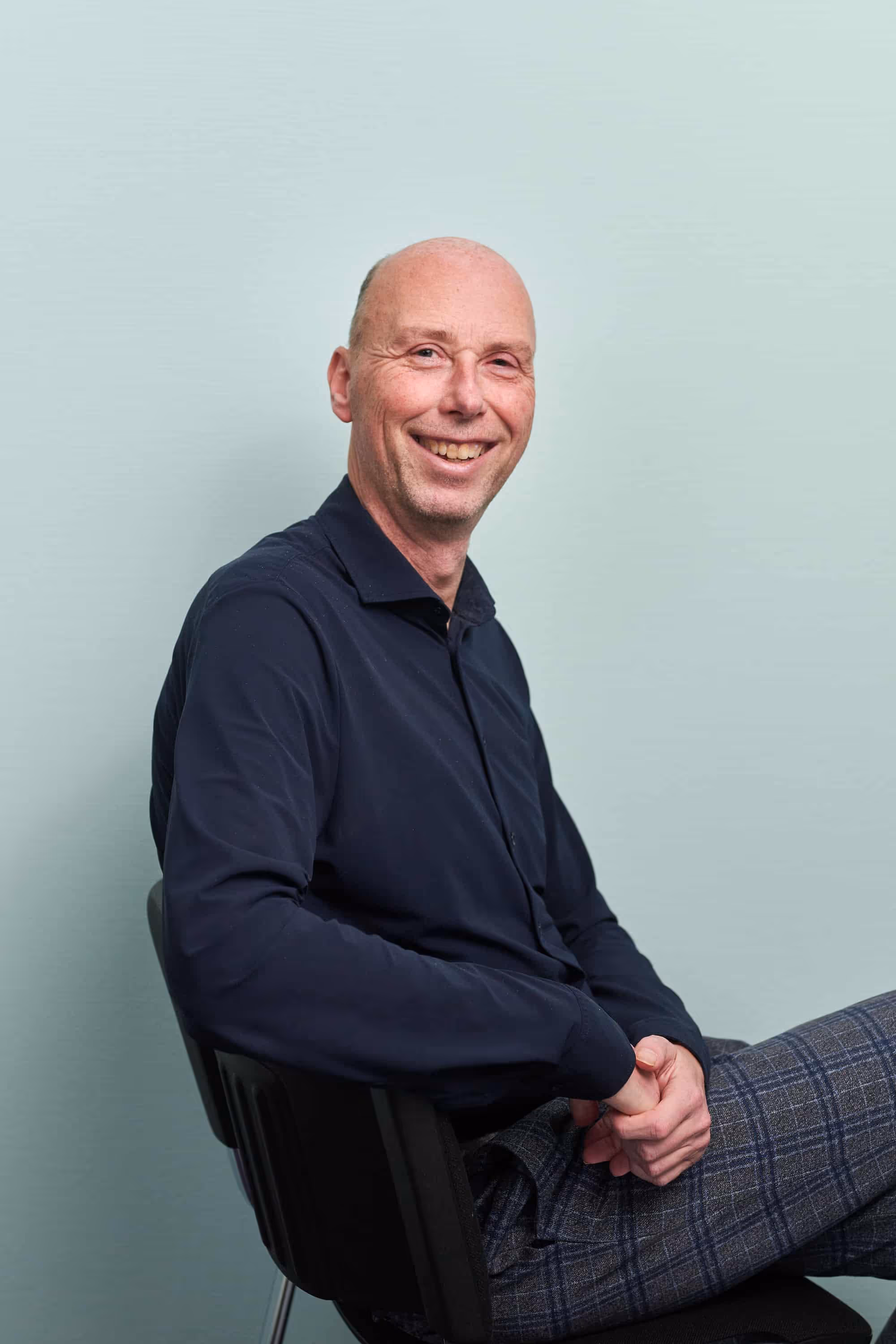 Lachende man met kale huid, zittend op een stoel, gekleed in een donkerblauw overhemd en geruite broek.
