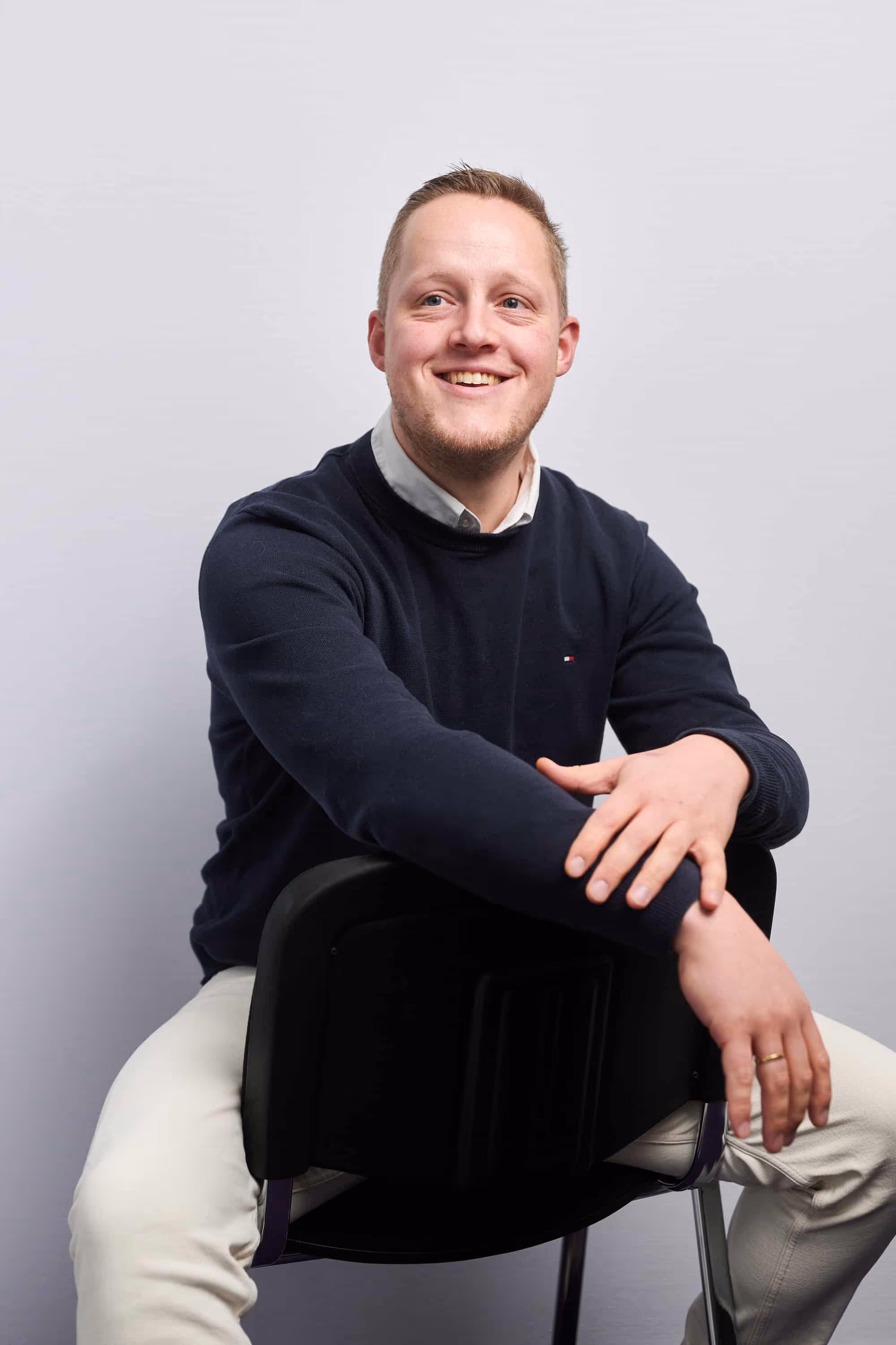Man in donkerblauwe trui en witte broek zit met armen over de rugleuning van een stoel, glimlachend naar rechts kijkend.