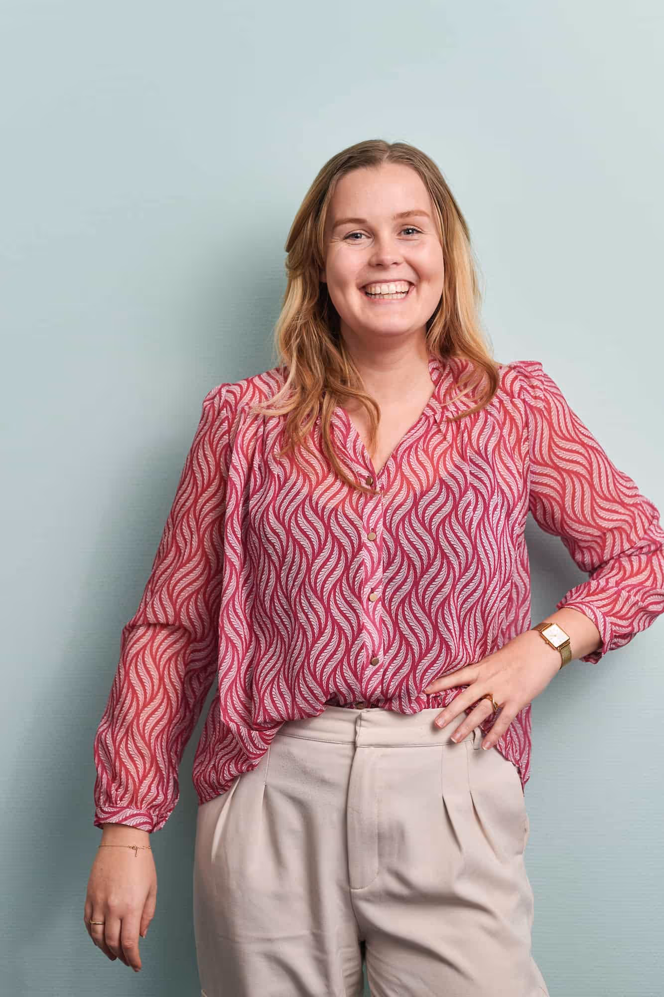Vrolijke vrouw met los haar draagt een roze blouse met patroon en beige broek, poseert tegen een lichtblauwe muur.