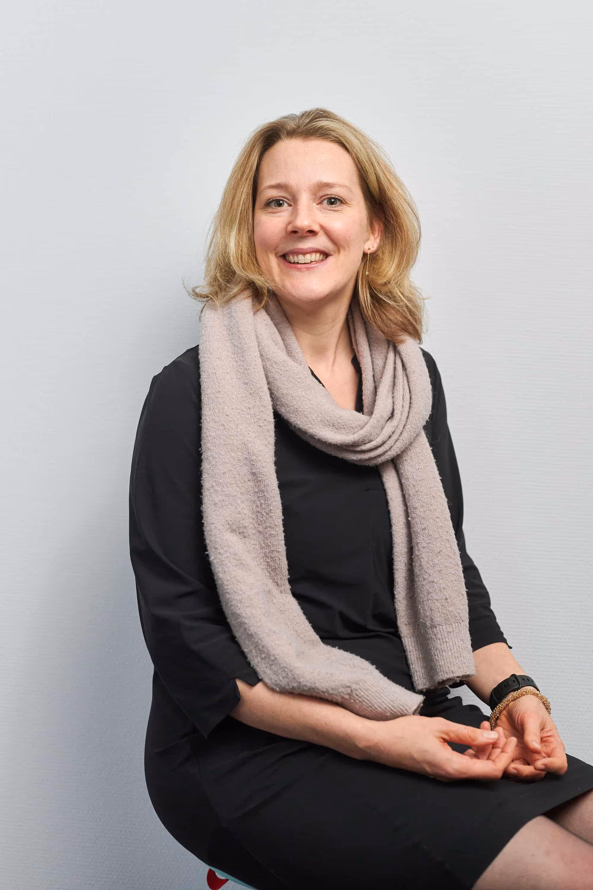 Vrouw met een lichte sjaal om haar nek en een zwarte jurk, zittend en glimlachend tegen een lichte achtergrond.