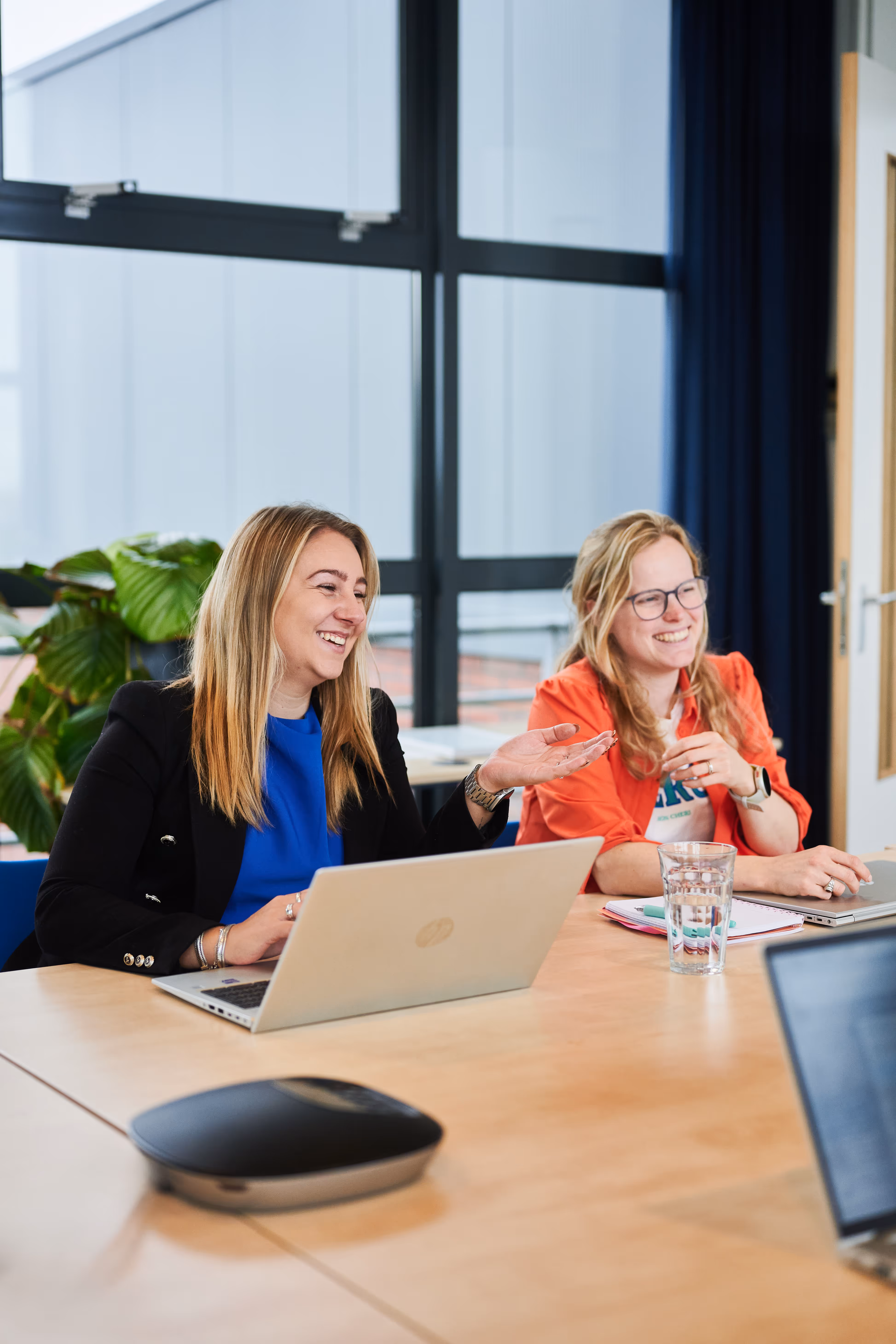 Twee vrouwen lachen en praten tijdens een vergadering aan een tafel met laptops en een glas water.