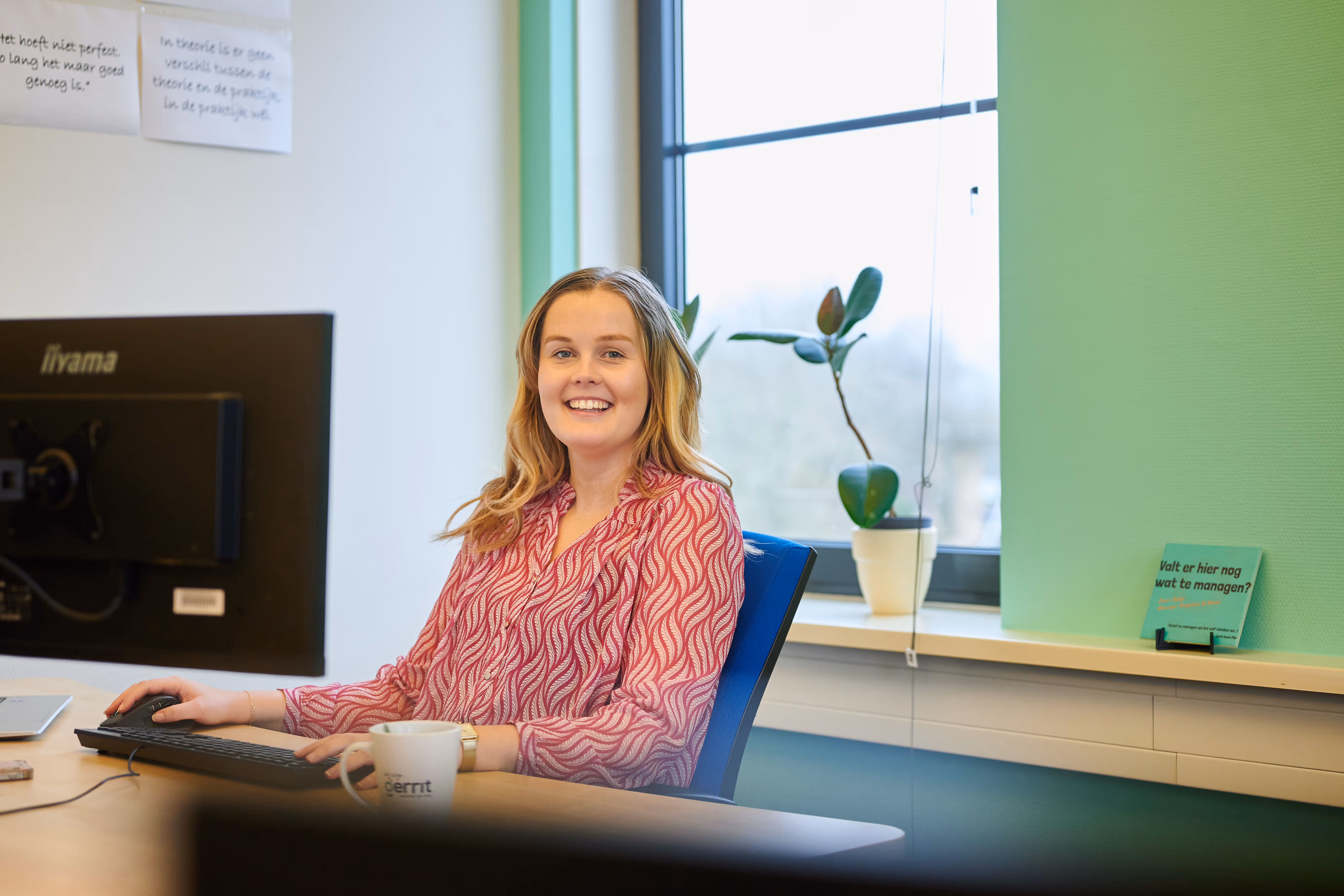 Jonge meid met kleurige blouse glimlacht naar camera vanachter de computer