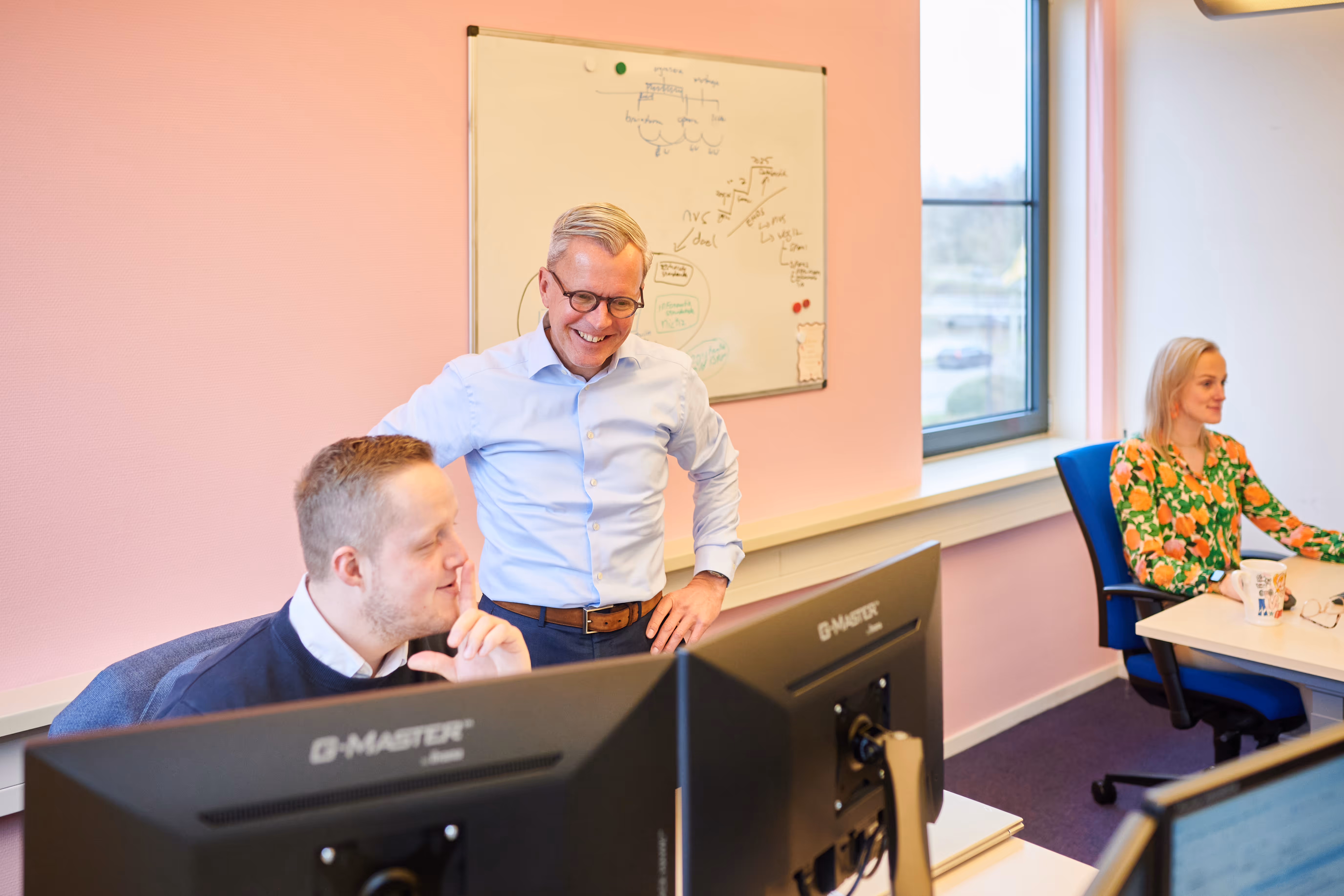 jonge man kijkt naar computerscherm en zit achter zijn bureau en oudere man staat erbij en kijkt mee aan de linkerkant, rechts zit een vrouwelijke collega achter haar computerscherm