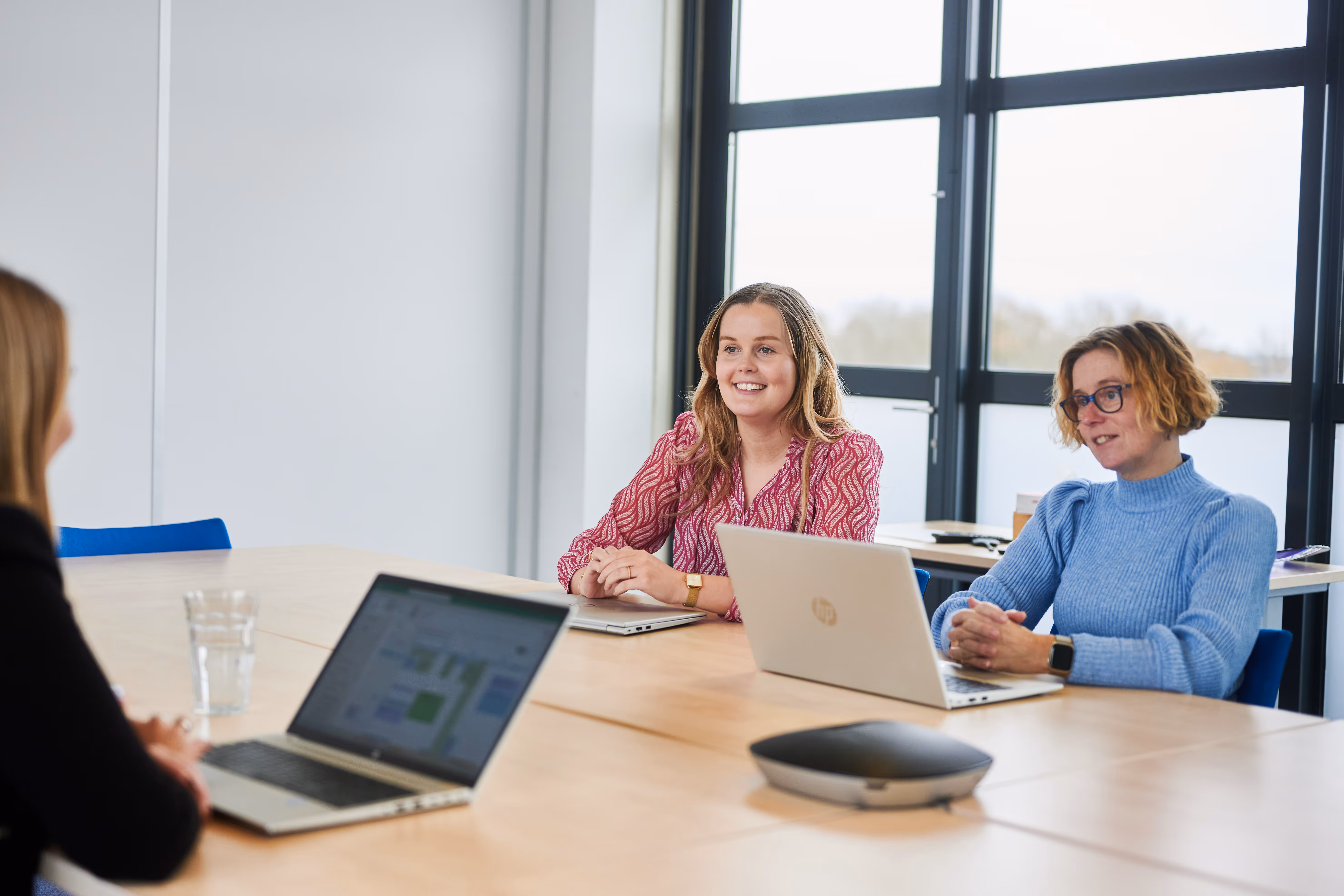 Drie collega's aan een tafel in overleg, één zit met de rug naar het beeld toe en heeft een laptop