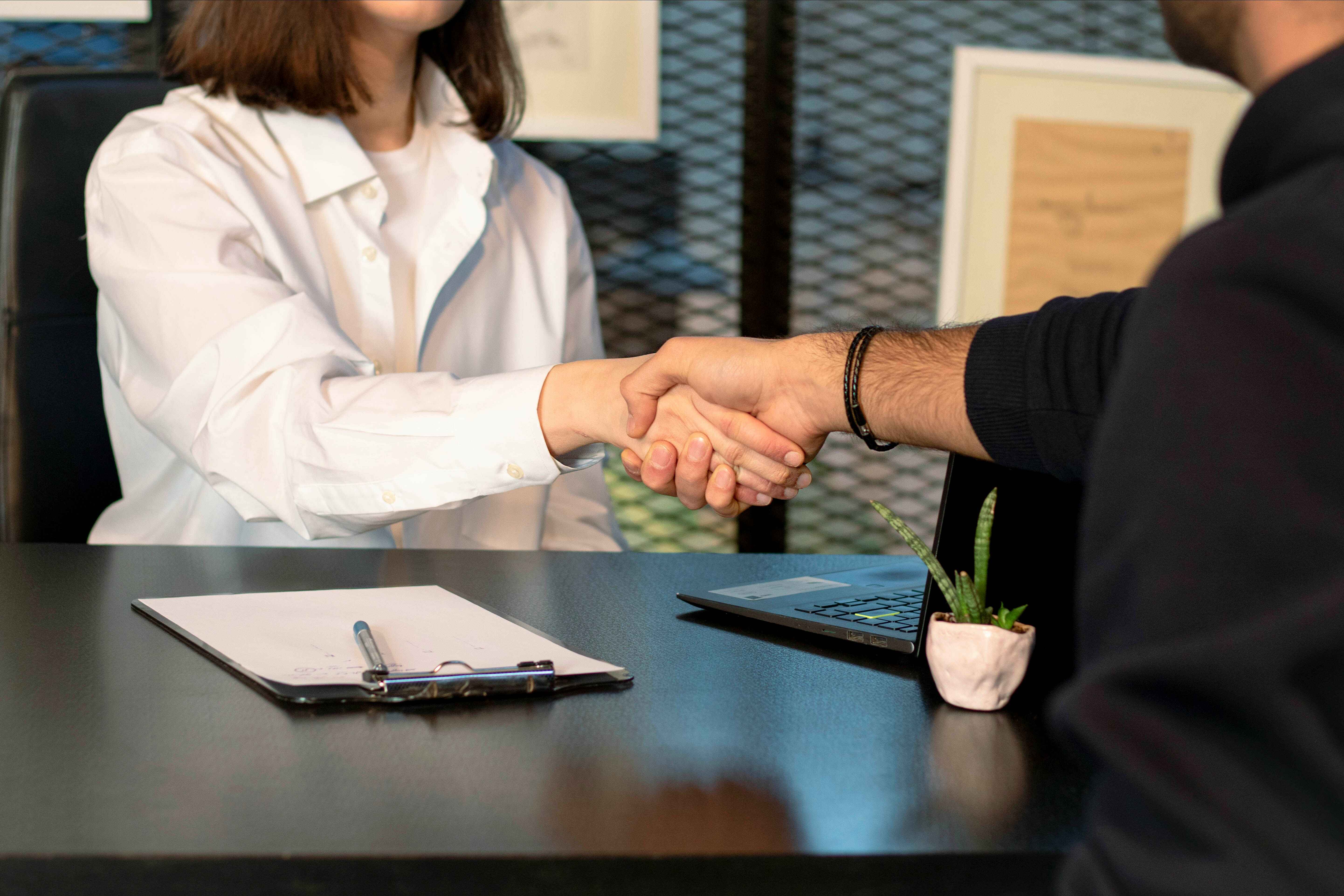 Two people shaking hands over a desk with a clipboard, pen, laptop, and small potted plant.