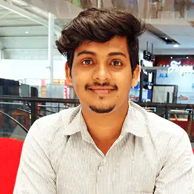 Young man with dark hair and a trimmed beard wearing a light-colored striped shirt, smiling indoors with a blurred background.