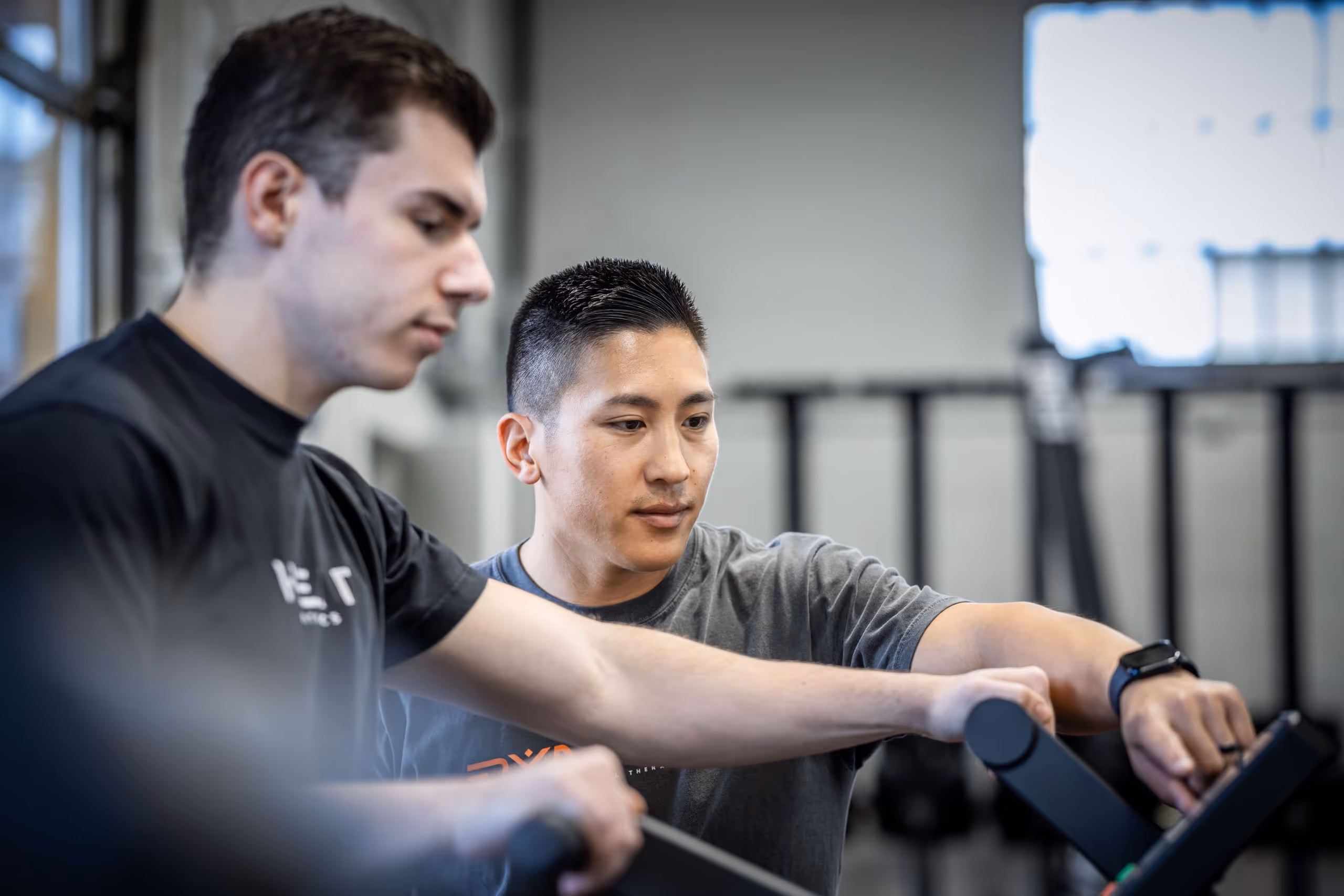 Two men focused on adjusting handles of gym equipment in a fitness center. Matt Chan massapequa physical therapy