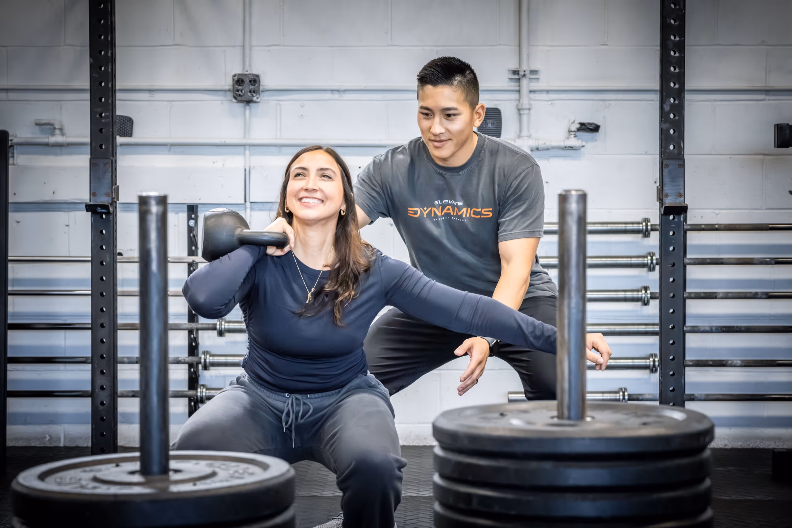 Woman squatting with a kettlebell resting on her shoulder while a trainer spots her in a gym. massapequa physical therapy