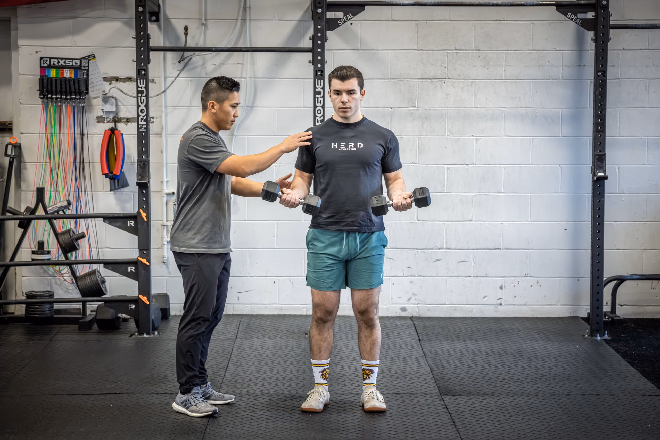 Trainer guiding a man lifting dumbbells while standing in a gym with exercise equipment in the background. physical therapy massapequa ny