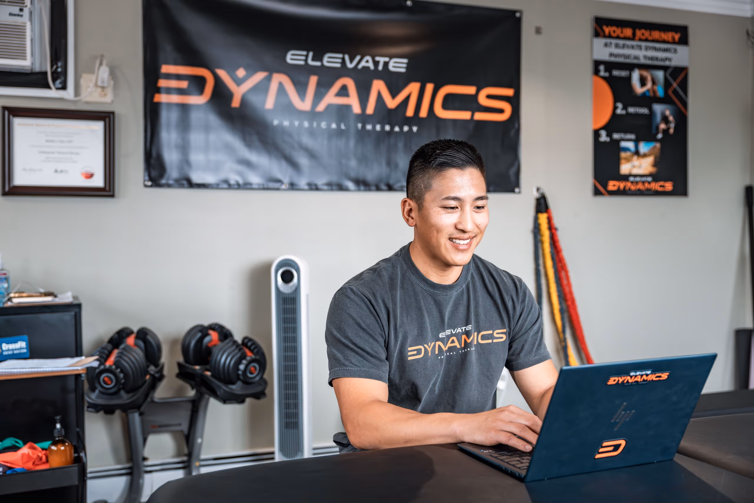 Matt Chan wearing an Elevate Dynamics Physical Therapy shirt working on a laptop in a gym setting. 
