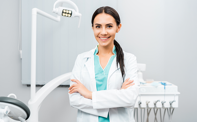 Smiling dentist standing in a modern dental office with dental equipment, providing family dentistry, preventive dental care, cosmetic dentistry, dental exams, teeth cleanings, crowns, fillings, dental implants, and restorative dental treatments.