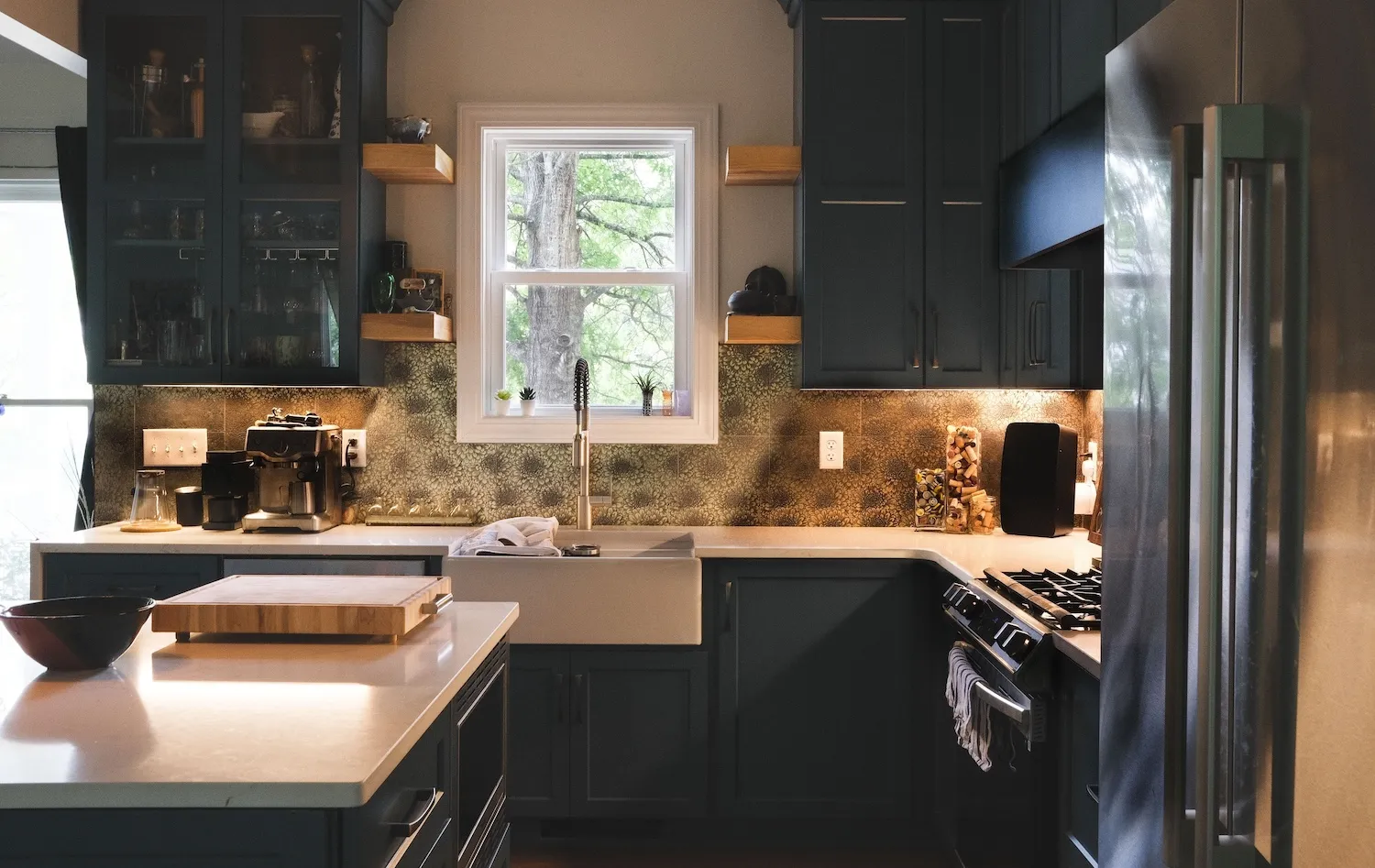 Modern kitchen with dark cabinetry, central island with cutting board and bowl, large window above farmhouse sink, coffee maker, and stainless steel refrigerator.