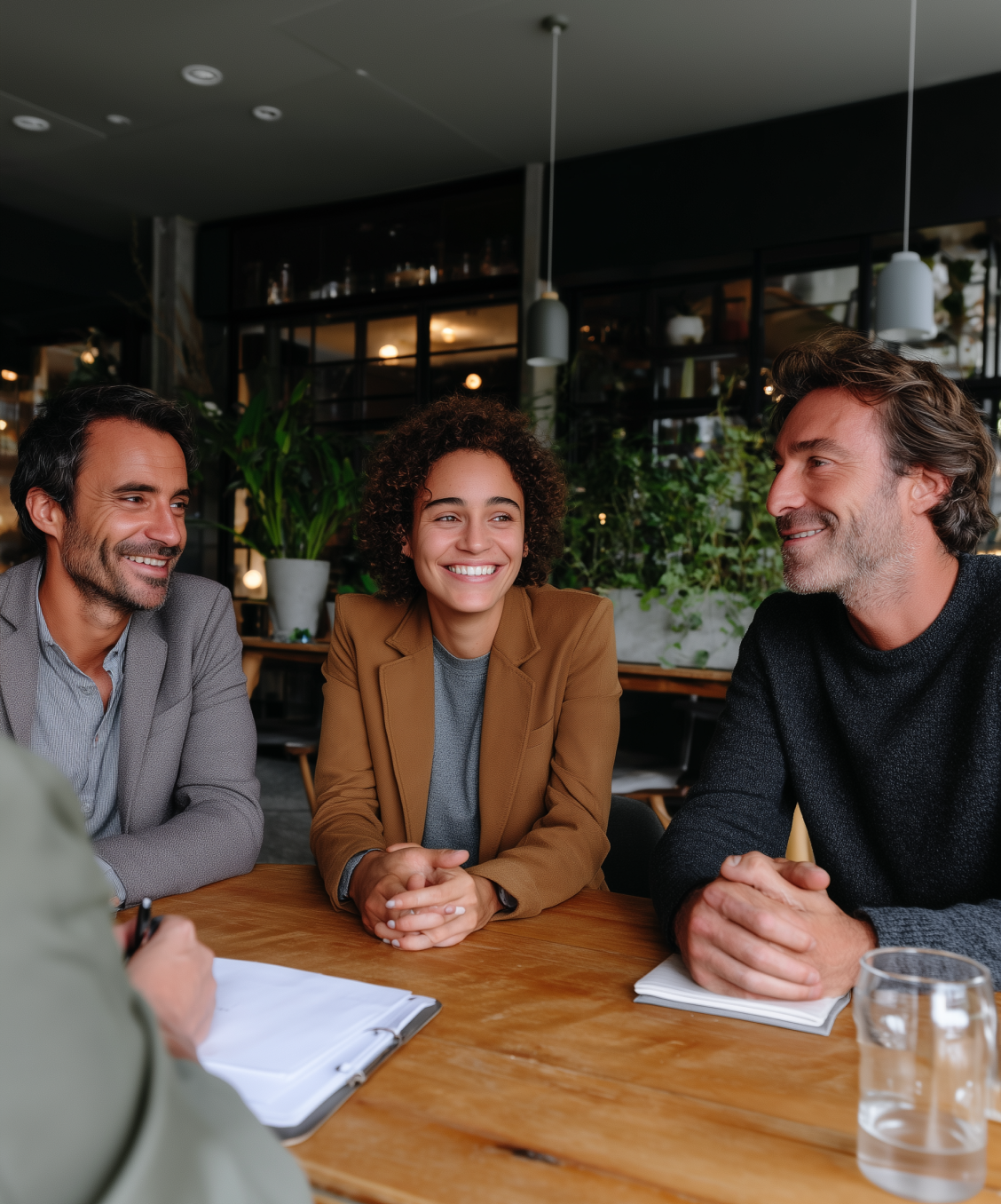 Three people sitting at a wooden table in a modern office, smiling and engaged in conversation.
