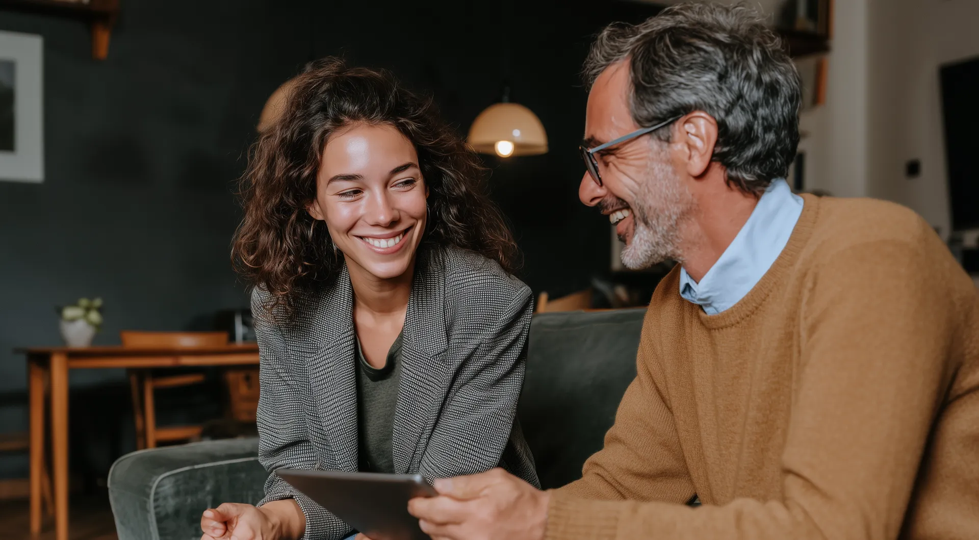 Smiling man and woman sitting on a couch, looking at a tablet and engaging in a friendly conversation.