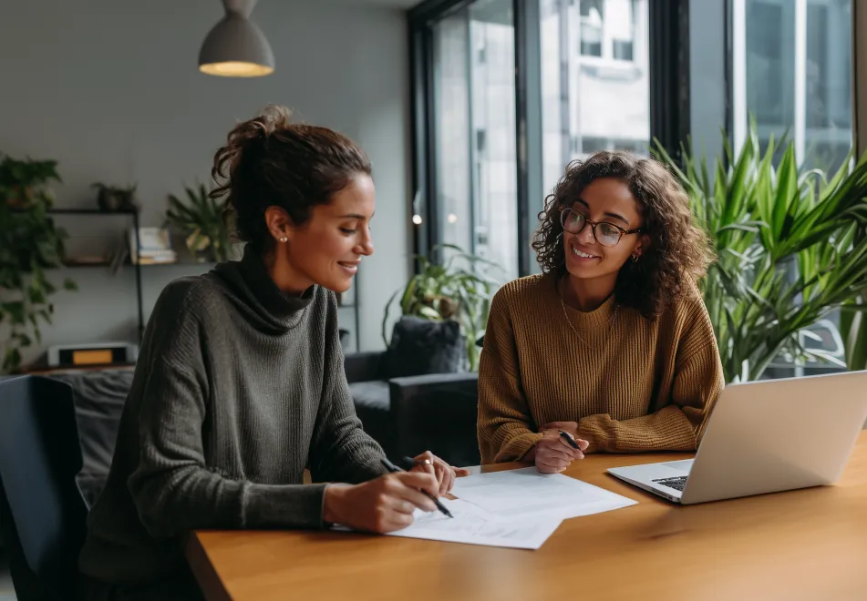 Two women sitting at a table with documents and a laptop, discussing work in a bright office with large windows and plants.