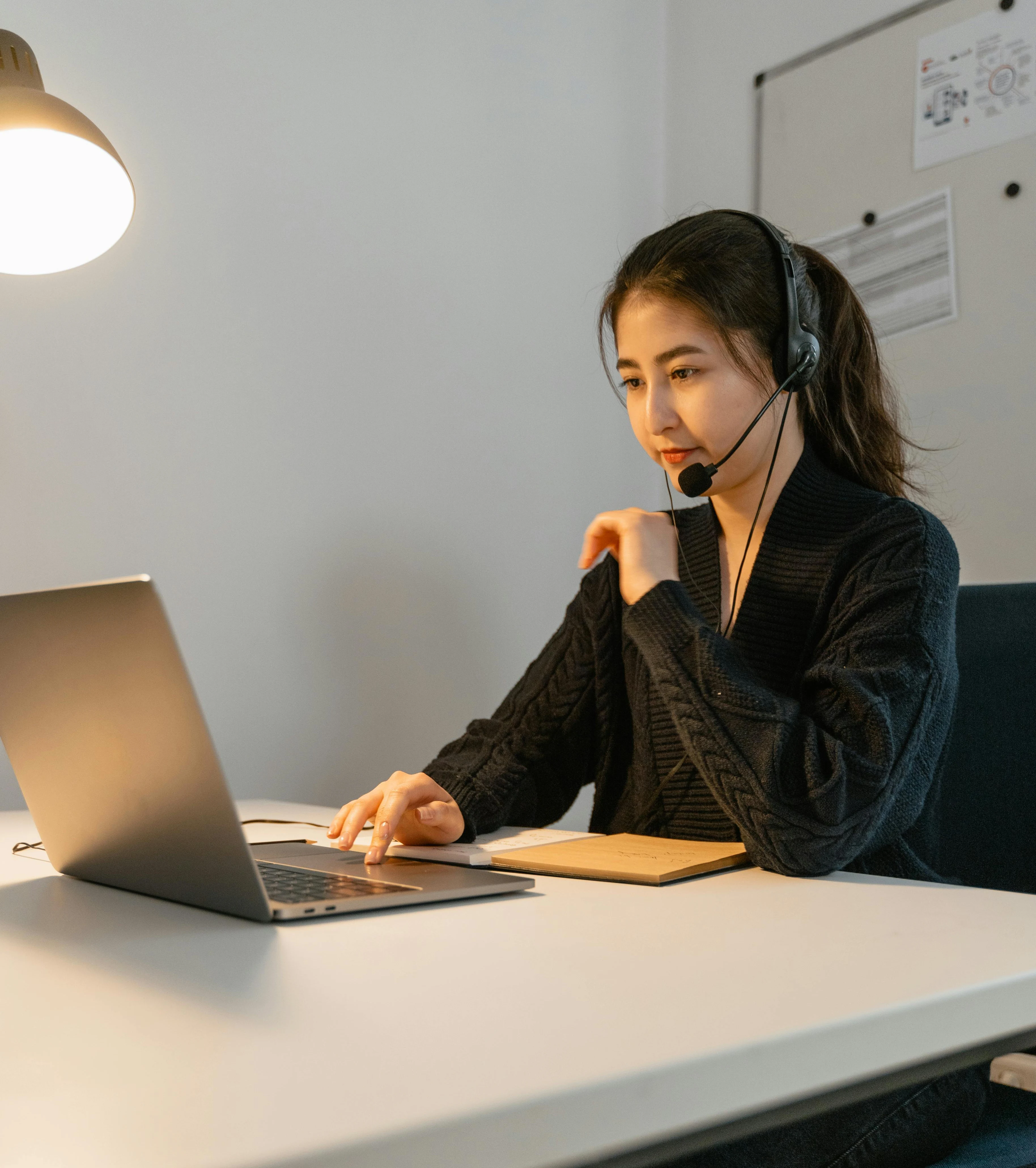 Woman wearing headset working on a laptop at a desk with a notebook, illuminated by a desk lamp.