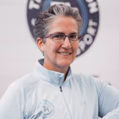 Smiling woman with short gray hair and glasses wearing a light blue zip-up jacket.