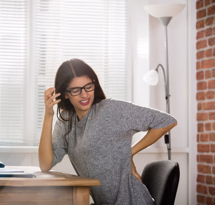 Young woman sitting at a desk in an office, holding her lower back in pain with a grimace.