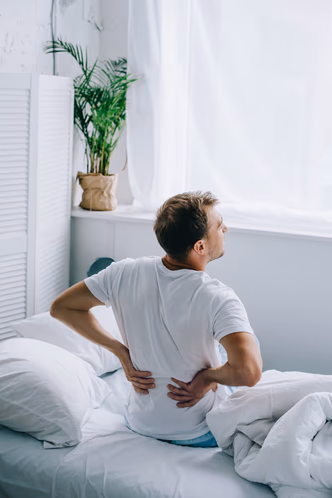 Man sitting on a bed holding his lower back in pain in a bright bedroom.