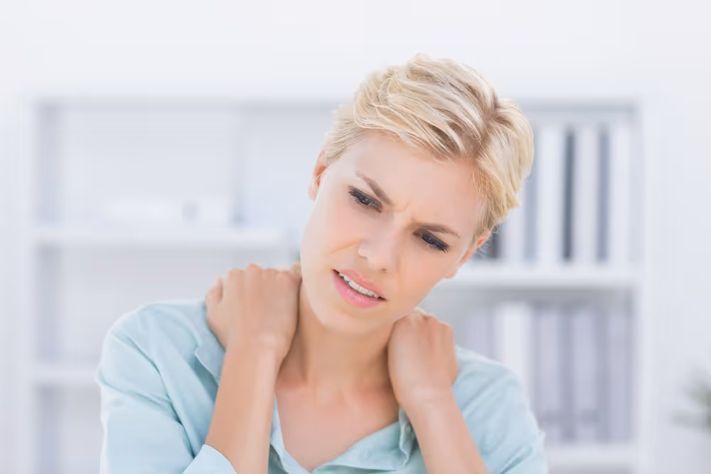 Woman with short blonde hair showing discomfort while holding her neck with both hands in a bright office setting.