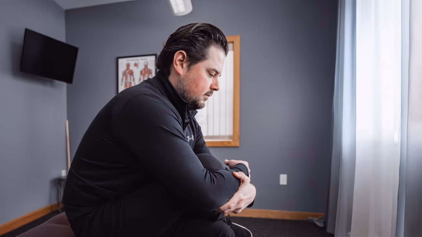 Man in black jacket sitting on a bench in a medical or therapy room, holding his elbow and looking down.