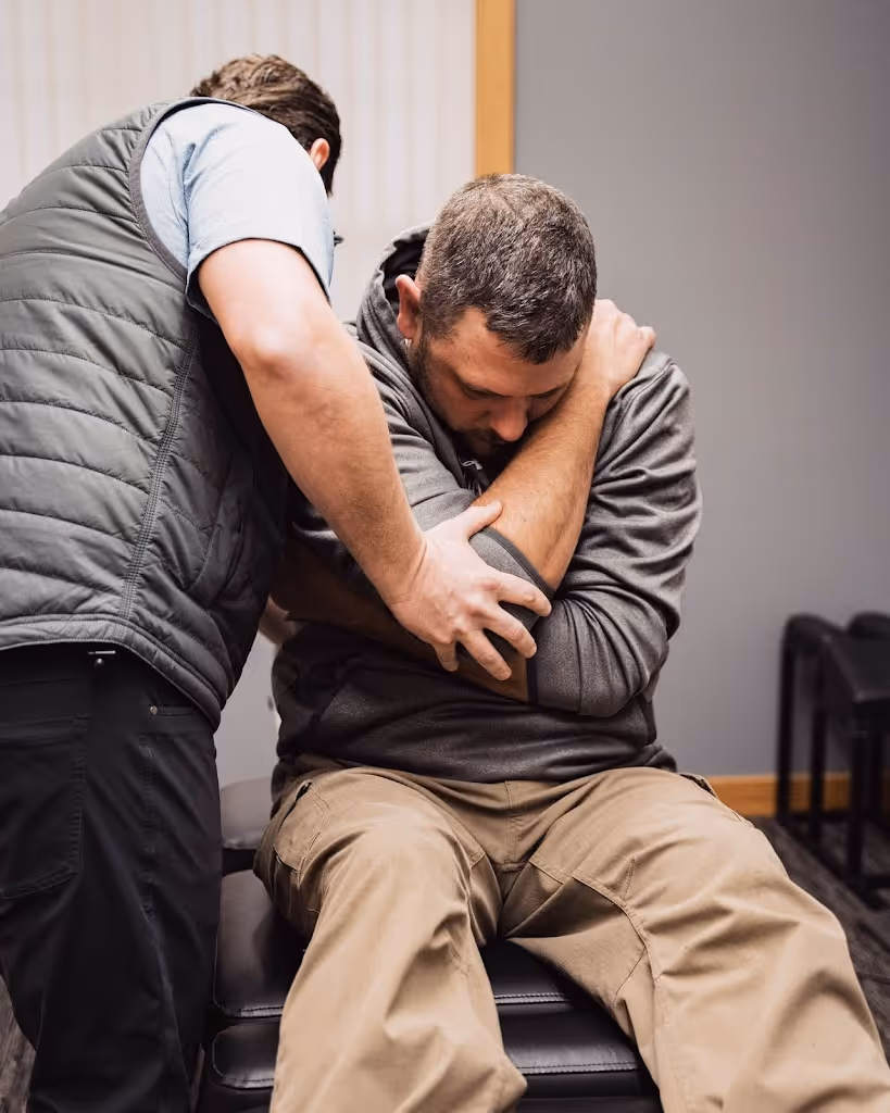 Chiropractor adjusting a seated man's shoulder and upper arm in a clinical setting.