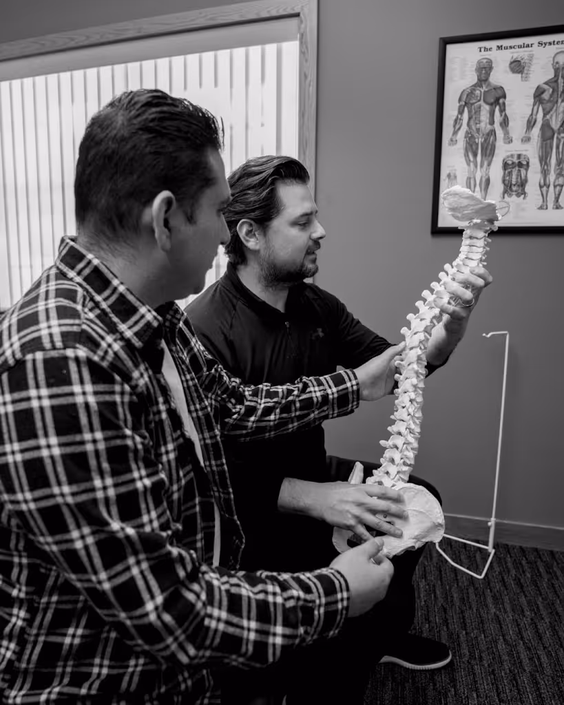 Two men examining a human spine model in a room with a muscular system chart on the wall.