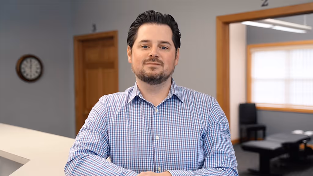 Man with dark hair and beard wearing a checkered shirt standing in an office with wooden doors and a clock on the wall.