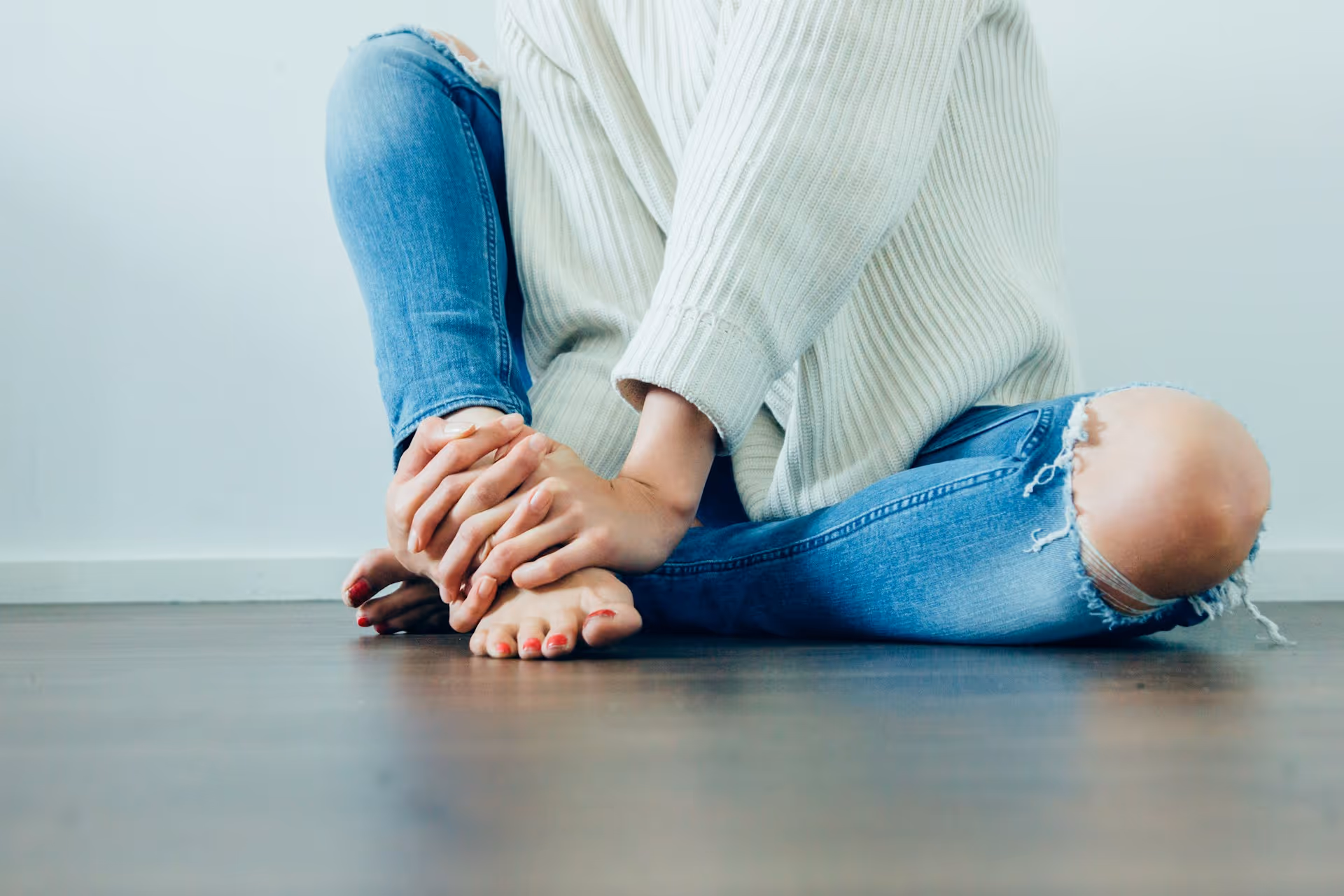 Person sitting on floor in ripped blue jeans and cream sweater, holding their foot with both hands.