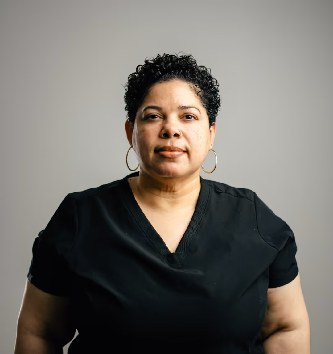 Woman with short curly hair wearing hoop earrings and a black V-neck shirt against a plain gray background.