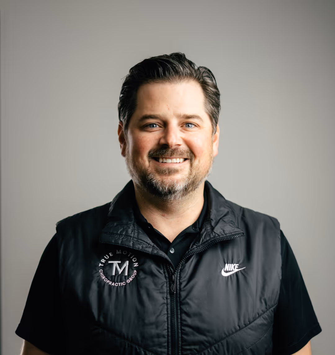 Smiling man with dark hair and beard wearing a black Nike jacket with True Motion Chiropractic Group logo on it against a plain light background.