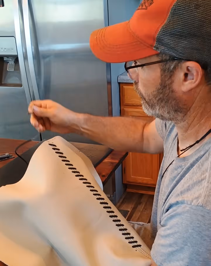 Man wearing glasses and orange cap stitching black thread into white material at a wooden table in a kitchen.