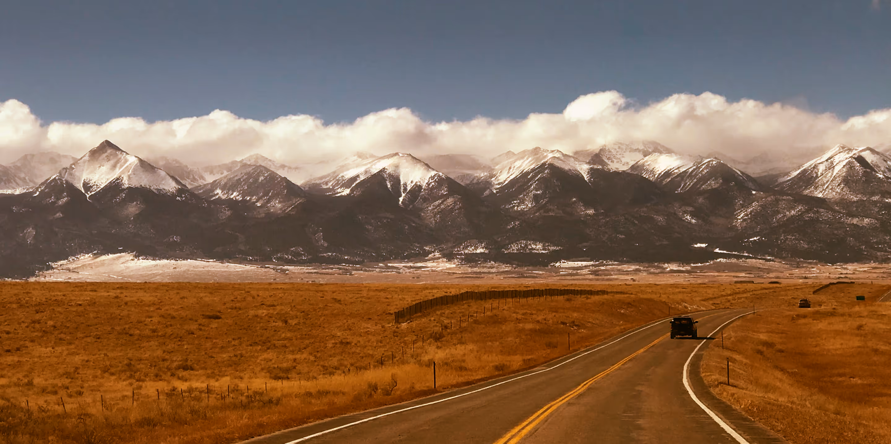 Two cars drive on a winding road through a golden field with snow-capped mountains and clouds in the background.