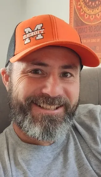Smiling man with a gray and black beard wearing an orange and black cap and a gray shirt, sitting on a gray couch with a colorful poster in the background.