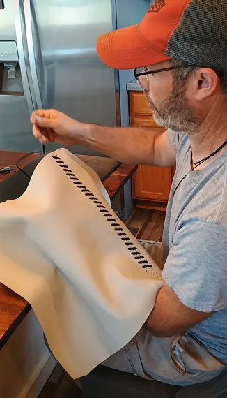Man wearing glasses and a cap hand-stitching black thread on a piece of beige leather at a wooden table.