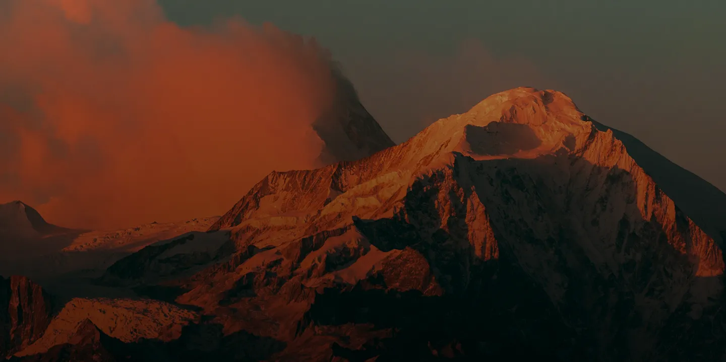 Mountain peaks illuminated by warm sunlight with orange clouds in the background during sunset.
