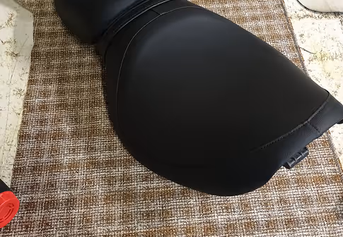Close-up of a black motorcycle seat resting on a textured brown carpet.