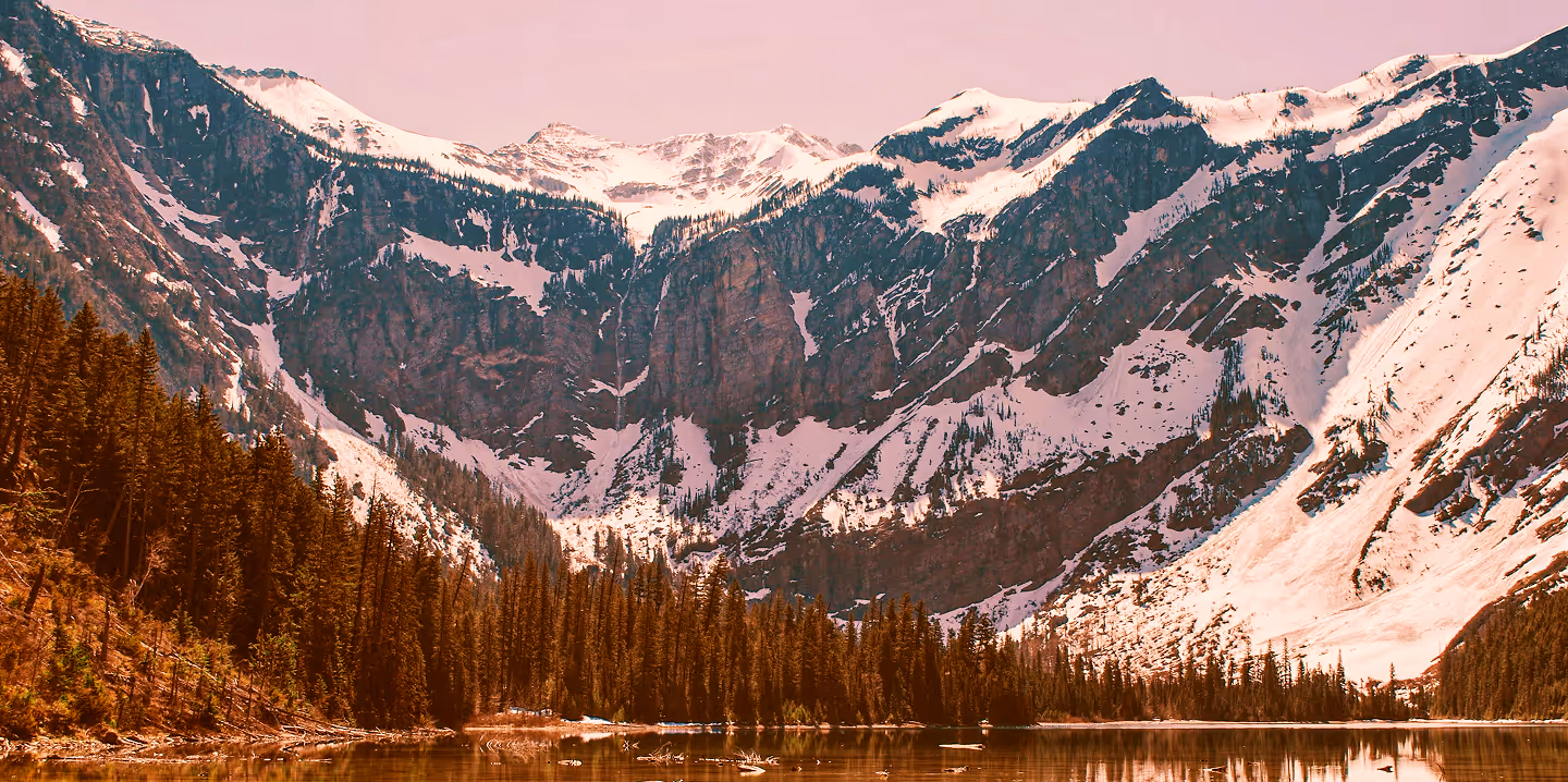 Snow-capped mountains behind a forest reflected in a calm lake under a pink sky.