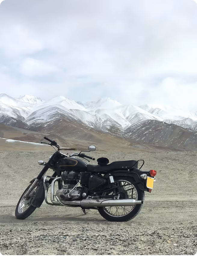 Black motorcycle parked on a gravel road with snow-covered mountains in the background under a cloudy sky.