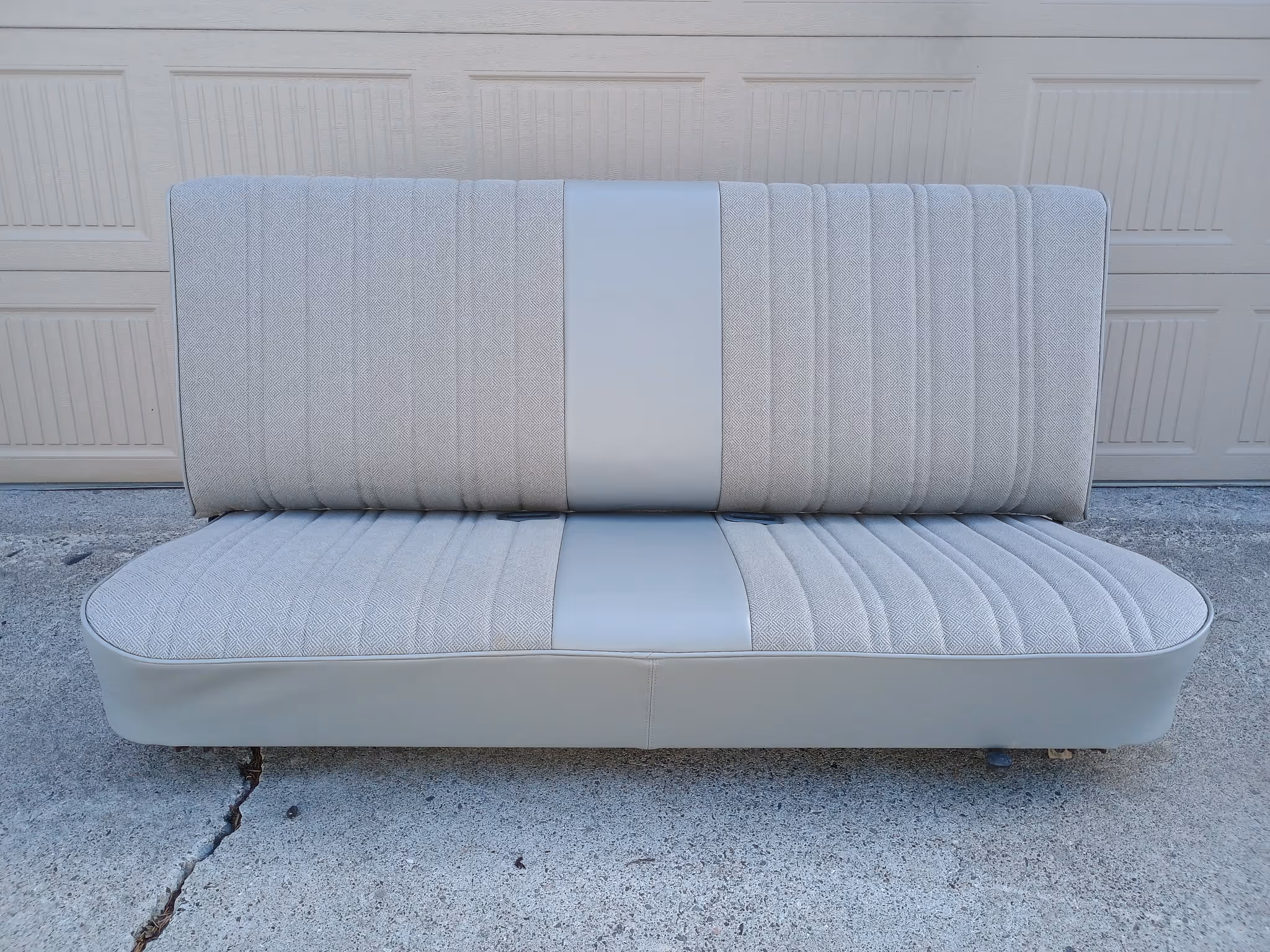 Light gray and beige two-tone vintage truck bench seat with textured fabric and vinyl inlay, placed on concrete in front of a garage door.