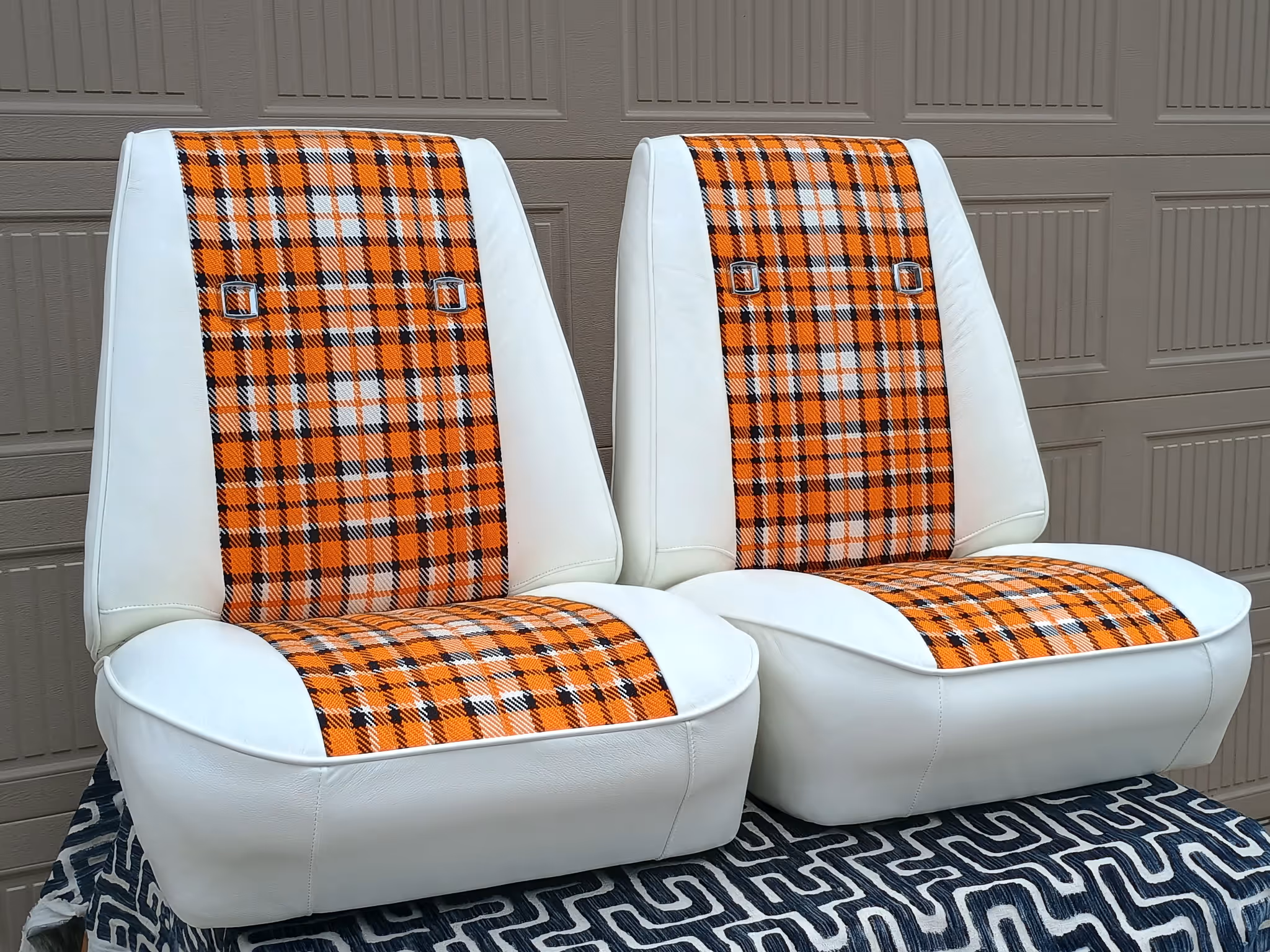 Pair of white bucket seats with orange and black plaid fabric inserts on seat backs and cushions, placed on a patterned cloth.