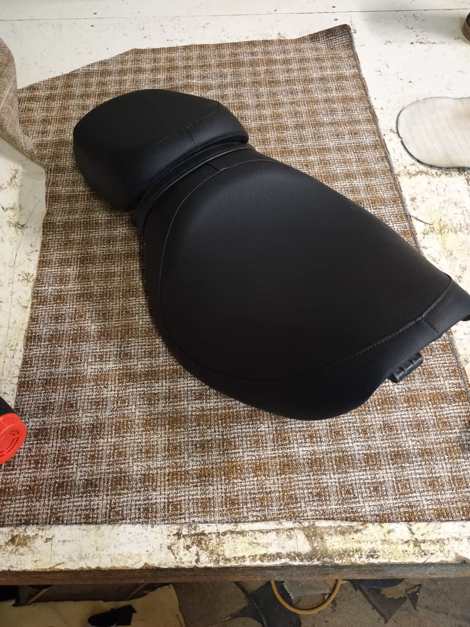 Black leather motorcycle seat resting on a brown and white patterned fabric on a workbench.