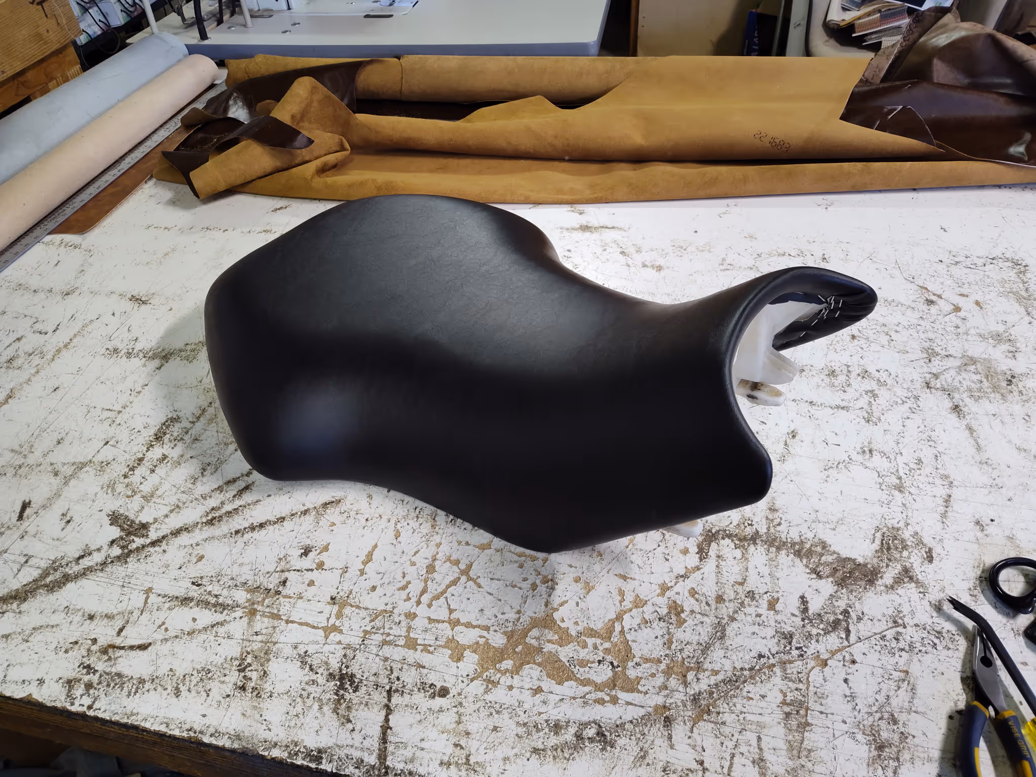 Black motorcycle seat resting on a worn, white workbench with upholstery materials and tools nearby.