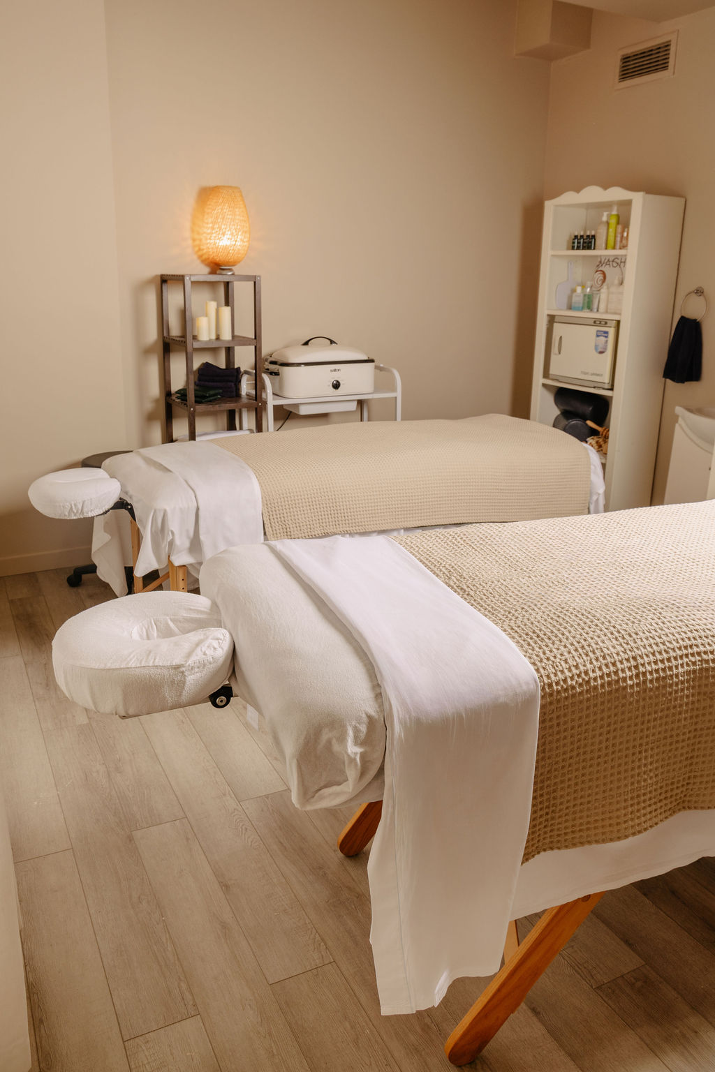 Serene massage room with two massage tables covered in beige and white linens, soft lighting, and shelves with towels and spa supplies.