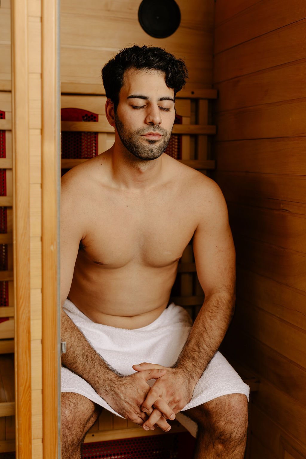 A shirtless man with a towel wrapped around his waist sits with eyes closed inside a wooden sauna.