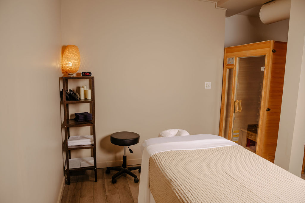 Quiet spa treatment room with massage table covered in beige blanket, wooden shelf with towels and candles, a stool, and a wooden sauna in the corner.