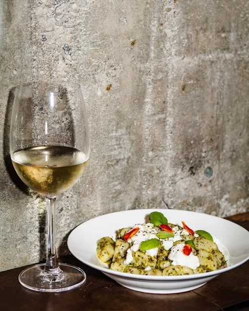 White wine in a glass next to a plate of gnocchi topped with green pesto, dollops of cream, red chili slices, and fresh basil leaves against a textured wall.