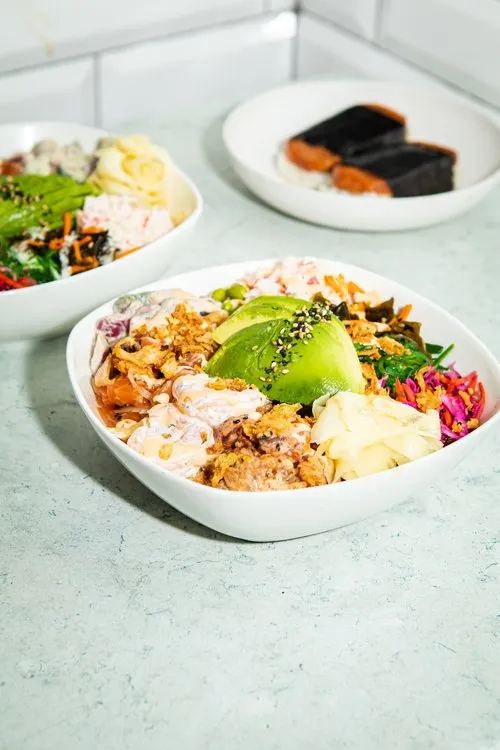 Three bowls of assorted poke with avocado, seaweed, greens, and shrimp on a light countertop.