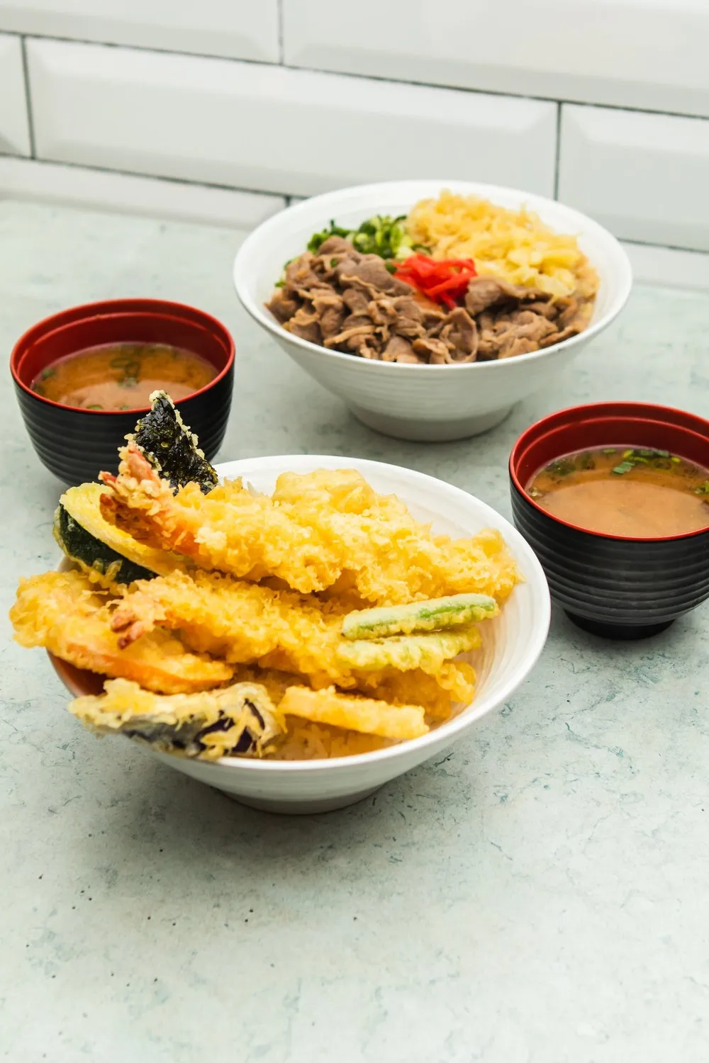 Two bowls with Japanese dishes: one with assorted tempura over rice and miso soup, the other with beef, pickled ginger, and vegetables with miso soup.